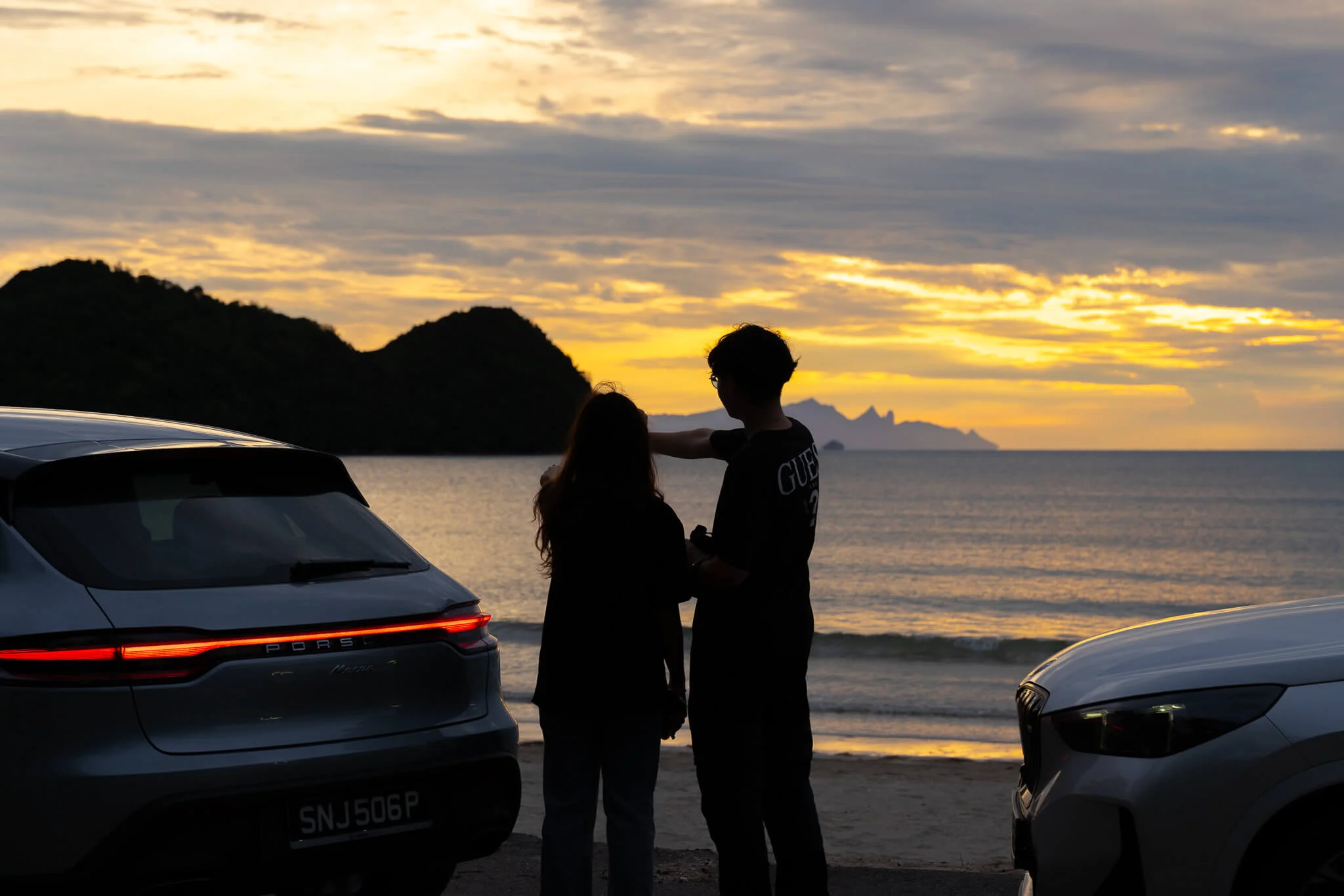 Couple by the beach with SUVs, sunset over the sea and islands.
